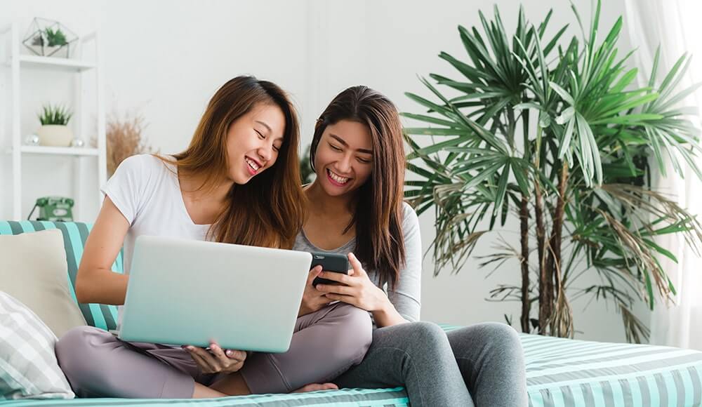 Two women laughing together while on the web on their laptop and smart phone